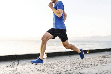 Foto de Musculoso corredor masculino corriendo terraplén en la mañana al amanecer, verano trotando a lo largo de la orilla del mar - Imagen libre de derechos