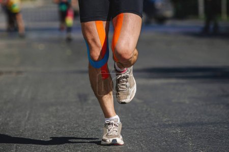 Téléchargez les photos : Jambes rapprochées coureur masculin courir asphalte sombre dans la course de marathon d'été, bande de kinesiotape sur les hanches et les genoux - en image libre de droit