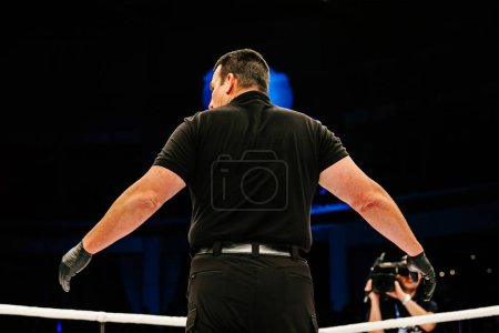 Téléchargez les photos : Vue arrière de l'arbitre debout dans l'anneau pendant le combat MMA sur fond sombre - en image libre de droit
