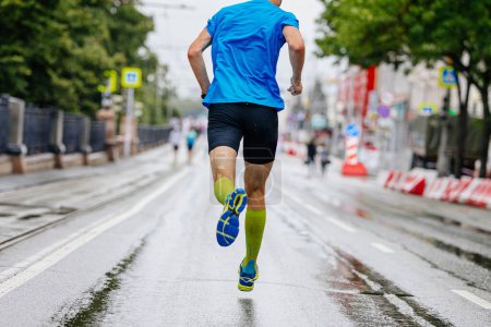 Foto de Vista trasera corredor masculino corriendo maratón de la ciudad en carretera mojada, calcetines de compresión en los pies, carrera deportiva de verano - Imagen libre de derechos