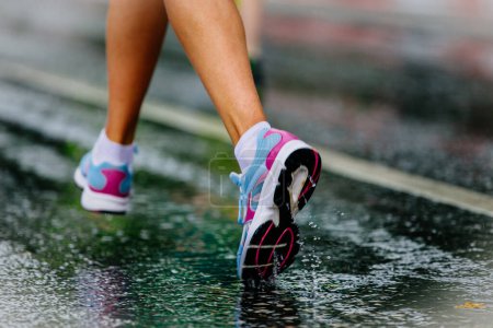 Foto de Primer plano parte piernas corredor femenino en zapatillas de correr correr sobre asfalto húmedo, gotas y salpicaduras de agua - Imagen libre de derechos