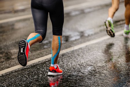 Téléchargez les photos : Jambes coureuse athlète coureuse course marathon sur asphalte mouillé, bande kinésio bleue sur les tibias - en image libre de droit
