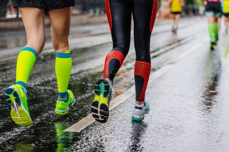 Téléchargez les photos : Jambes coureuses courir ensemble marathon sur asphalte humide, chaussettes de compression vertes et leggings noirs - en image libre de droit