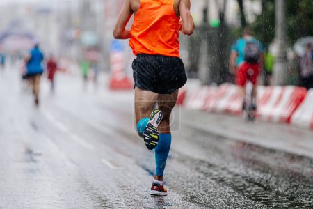 Photo for Rear view runner athlete run marathon race on rain, in blue compression sleeve on feet and orange singlet - Royalty Free Image