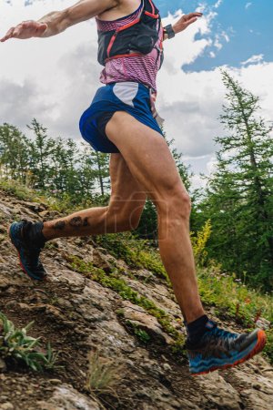 Foto de Primer plano corredor masculino corriendo por empinada ladera de la montaña, carrera de maratón sendero de verano - Imagen libre de derechos