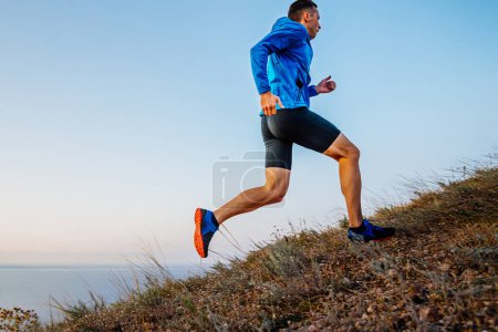 Foto de Vista lateral atleta corredor masculino corriendo cuesta arriba en chaqueta azul y medias negras, fondo del cielo y el mar - Imagen libre de derechos