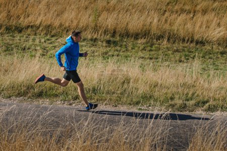 Photo for Middle-aged male runner in blue jacket running road in field of dry grass - Royalty Free Image