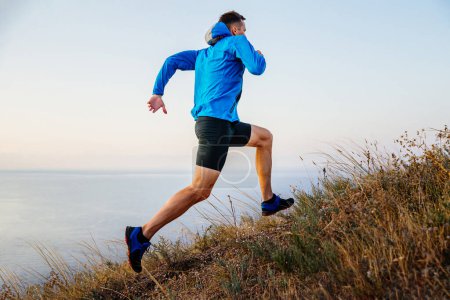 Photo for Male runner running uphill in blue jacket and black tights, background of sky and sea, trail dry grass - Royalty Free Image