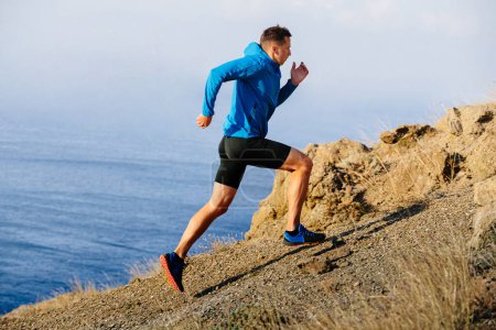 Téléchargez les photos : Athlète coureur en montée de montagne en veste bleue et collants noirs, fond de mer - en image libre de droit