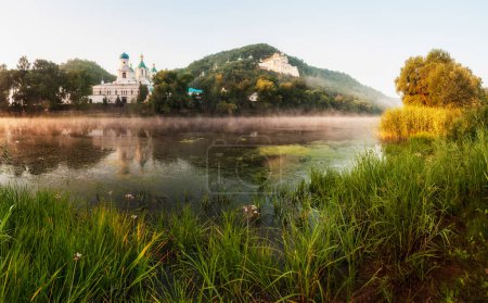 Téléchargez les photos : Belle matinée lever de soleil avec du brouillard sur l'eau. Temple sur la rive au lever du soleil, reflété dans la rivière. Temple sur des falaises de craie à l'aube, au-dessus de la rivière. Paysage ukrainien traditionnel. - en image libre de droit