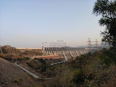 Téléchargez les photos : L'image montre un grand barrage avec une centrale électrique en arrière-plan, entouré d'une végétation luxuriante et un ciel bleu clair. - en image libre de droit