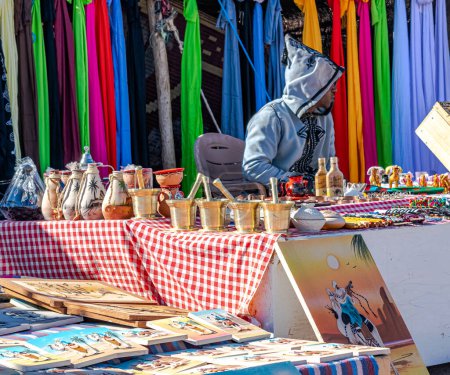 Téléchargez les photos : Exposition of souvenir products at a market of Sahara desert. Fabrics of many decorative objects and costume jewelry, mortar and pestle, vases, leather wallets and purses in the shape of babouche. - en image libre de droit