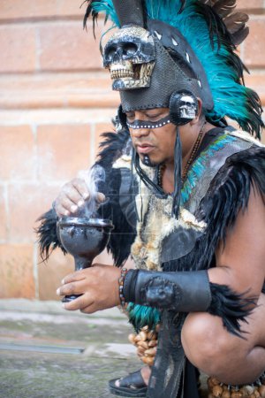 Photo for Close up of a concentrated shaman smelling smoke from a calyx or ritual chalice, aztec dancer with feathered headdress and skulls on his costume, hispanic culture mexican traditional performer - Royalty Free Image