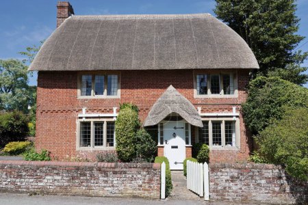Exterior view and garden of a beautiful old thatched roof red brick cottage house on a quiet street in an English village 
