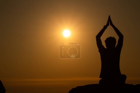 Foto de Silueta del Joven haciendo yoga o meditación u orando al borde de una montaña al atardecer, Día Mundial del Yoga - Imagen libre de derechos