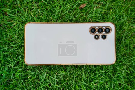 White phone with gold frame and multiple cameras on a green grass during hot sunny day.