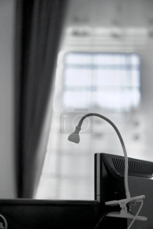 A white minimalist clip-lamp placed on toop of a computer monitor screen to give a top-down (semi spotlight) light feeling for the user. Photographed during the day, with a softbox-feeling light comes from the window on the background.
