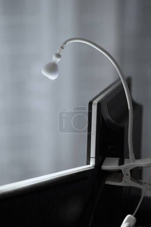 A white minimalist clip-lamp placed on toop of a computer monitor screen to give a top-down (semi spotlight) light feeling for the user.