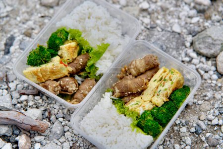 Köstliche Rindfleisch-Enoki-Rolle, Omelette, gedämpfte Brocolli und Reis in einer Lunchbox aus Plastik. Im Hintergrund wurde ein weiß-gelb-schwefelhaltiger Kiesboden gezeigt.
