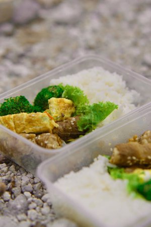Köstliche Rindfleisch-Enoki-Rolle, Omelette, gedämpfte Brocolli und Reis in einer Lunchbox aus Plastik. Im Hintergrund wurde ein weiß-gelb-schwefelhaltiger Kiesboden gezeigt.