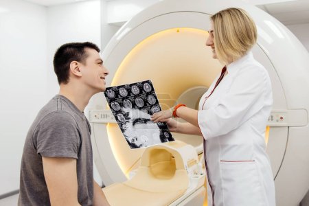Foto de Radióloga doctora que explica los buenos resultados de la tomografía computarizada al hombre joven que muestra rayos X con imágenes, observando y analizando la tomografía computarizada en una clínica moderna junto al tomógrafo moderno - Imagen libre de derechos