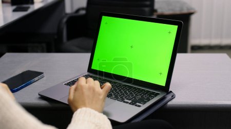 Téléchargez les photos : Par-dessus l'épaule d'une femme d'affaires qui travaille à l'intérieur du bureau sur PC sur le bureau, en regardant l'écran vert. Personne de bureau utilisant un ordinateur portable avec écran vert pour ordinateur portable, assise à une table en bois - en image libre de droit