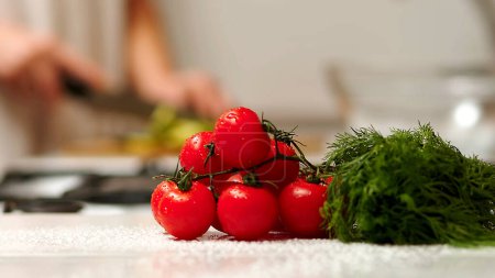 Frühlingssalat mit Tomaten, Gurken, Gemüse und Mischungen. Frau schneidet Gurke für Salat auf Holzschneidebrett auf Küchentisch mit Gurken, Tomatenzweig und Schalotte herum.