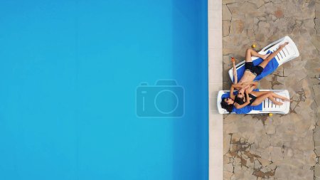 Téléchargez les photos : Un couple amoureux, un homme et une jeune femme, s'allongent sur des chaises longues près de la piscine, profitent de vacances tropicales et de la compagnie l'un de l'autre. Vue d'en haut. - en image libre de droit