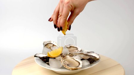 Téléchargez les photos : Huîtres dans une assiette ronde avec glace, citron, la main d'une femme verse un citron ouvert sur les huîtres. restaurant de fruits de mer - en image libre de droit