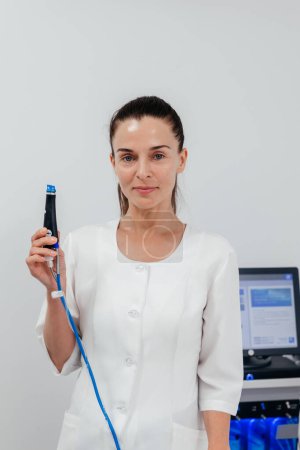 Middle aged beautician holding ultrasound device for face lifting and skin tightening procedure. Young woman cosmetologist using modern cosmetology equipment in clinic