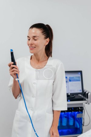 Middle aged beautician holding ultrasound device for face lifting and skin tightening procedure. Young woman cosmetologist using modern cosmetology equipment in clinic