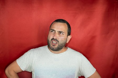 Hispanic young adult man thoughtful looking up indecisive and confused with idea in red background