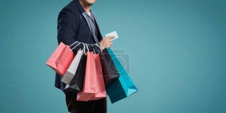 Photo for Cropped image of a man in a suit holding shopping bags and a credit card on a teal background - Royalty Free Image