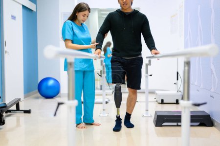 Téléchargez les photos : Physiothérapeute aidant le patient avec prothèse jambe marcher entre les barres parallèles pendant la séance de physiothérapie à l'hôpital, Nouvelle production de membres artificiels pour les personnes handicapées - en image libre de droit
