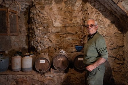 Photo for Authentic older man, dressed in camouflage, with sunglasses, inside an old wine cellar of a village house in the rocky interior, barrels and demijohns in the background. - Royalty Free Image
