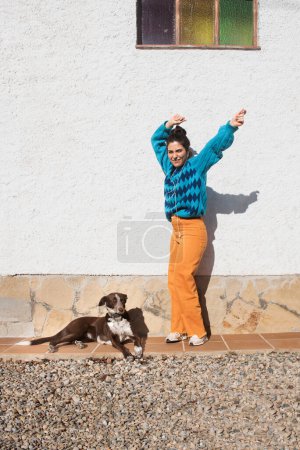 Photo for Hunting dog and young latin girl in colorful clothes, saturated blue argyle sweater and orange pants, white stucco wall background outside, light sunny day. Listen music with headphones and dancing - Royalty Free Image