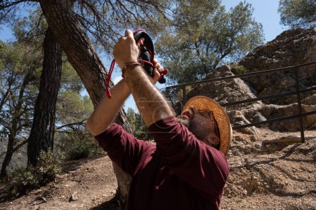 man drinking from a wineskin, wearing an explorer's cap and a maroon T-shirt in a natural setting