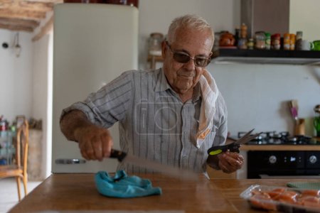 achtzigjähriger Mann in der Küche mit einem Lappen auf der Schulter und einem Brotmesser, der das Hackholz mit einem Holzlöffel säubert.