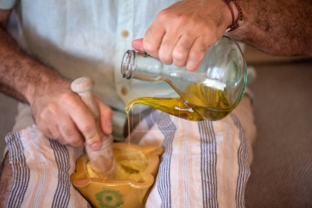 Téléchargez les photos : Homme en short avec chiffon sur les jambes faisant "allioli". une sauce typique à l'ail et à l'huile de Catalogne, il mélange la sauce dans un mortier traditionnel avec un pilon en bois et verse l'huile dans l'huile d'olive. - en image libre de droit