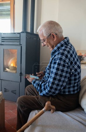 Téléchargez les photos : Vieil homme de quatre-vingts ans regardant l'écran de téléphone portable, assis sur le canapé à côté de la cuisinière à pellets. Pantalon en velours côtelé marron et chemise en flanelle à carreaux bleue. - en image libre de droit