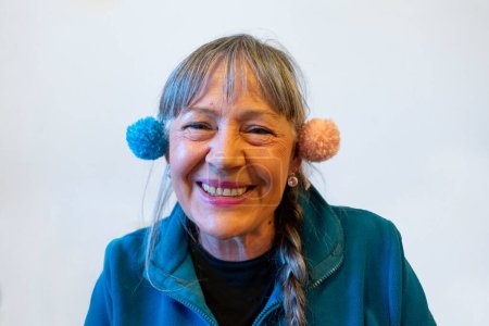 Frau mit Pompons über den Ohren lacht. Person um die sechzig mit langen grauen Haaren und langem Zopf. Gekleidet in ein blaues Vlies. Klarer Hintergrund
