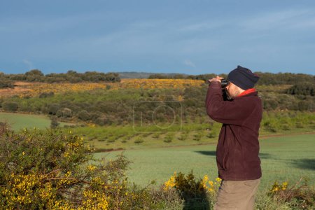 Man in profile looking through binoculars, dressed in mountain clothes, brown trousers, maroon fleece and neckerchief on his head. Field full of flowering gorse