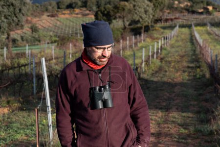 man in the field with binoculars hanging down, dressed in mountain clothes, brown trousers, maroon fleece and neckerchief on his head. Behind field of vines