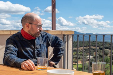hombre de cuarenta años en una terraza mirando al paisaje, apoyado en una mesa con una cerveza y un plato de aperitivo, afuera con una vista de la naturaleza, hombre calvo, con gafas, una camisa a cuadros y una bufanda alrededor de su cuello, estilo montañero.
