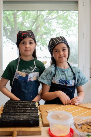 Zwillingsschwestern posieren nach der Herstellung von Sushi-Rollen. In Jeansschürze und schwarzer Kochmütze gekleidet, lächelnd in die Kamera schauend.
