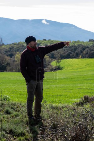 Man in landscape of sown fields looking to one side and pointing with his arm, dressed in polar jumper and polar cap, mountaineer style.