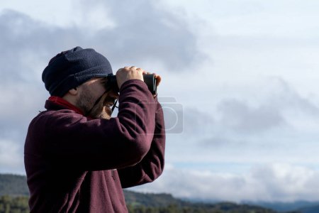 Man in his forties looking through binoculars in mountain clothes, with glasses and beard, exploring the landscape.