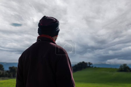 Man in a landscape of sown fields looking to the side, dressed in a polar jumper and polar cap, mountaineer style.