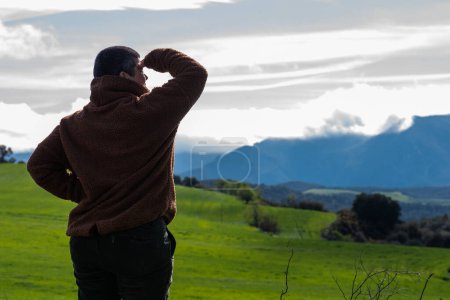 woman shielding her eyes from the sun looking at a landscape of sown fields, dressed in a polar jumper and short hair.