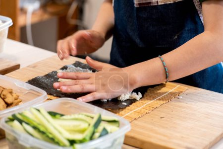 Photo for Detail of girl's hands making sushi, squeezing rice on nori seaweed, in front of lunch box with sliced cucumber pieces ready for the little girl to cook on her own - Royalty Free Image